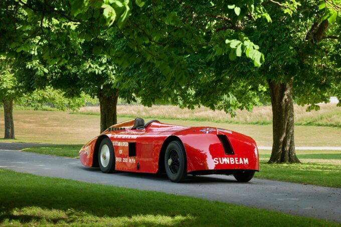 Restored red Sunbeam 1000hp land-speed car parked beneath trees at Beaulieu, seen from the rear quarter