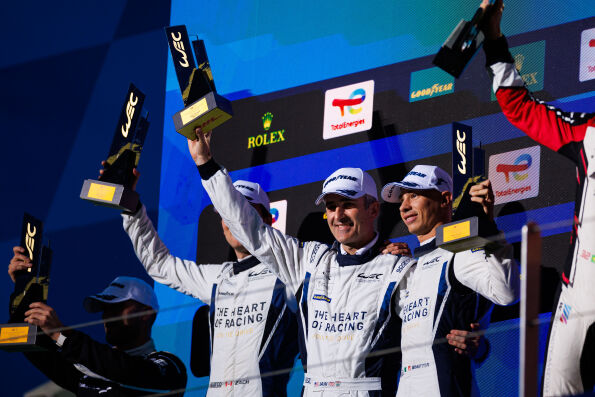 The Heart of Racing Team drivers raise trophies on the LMGT3 podium at Fuji Speedway.