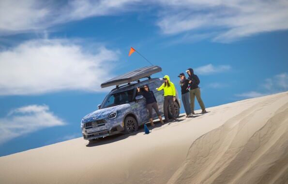 MINI's Rebelle Rally team beside a camouflaged MINI SUV on a sand dune during training.
