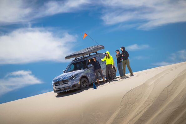 MINI's Rebelle Rally team beside a camouflaged MINI SUV on a sand dune during training.