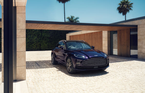 Front three-quarter view of a dark purple Aston Martin DBX GT parked in front of a modern stone and timber house with palm trees