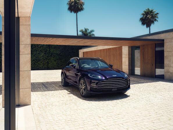 Front three-quarter view of a dark purple Aston Martin DBX GT parked in front of a modern stone and timber house with palm trees