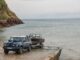 Blue INEOS Grenadier Helmsman recovering a motorboat on a trailer at a coastal slipway